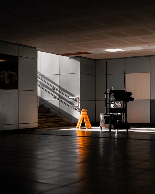 A dimly lit indoor space featuring a tiled floor and a backup cleaning cart with supplies, positioned near a staircase with metal handrails and a beige wall. The scene shows sunlight streaming in, cre